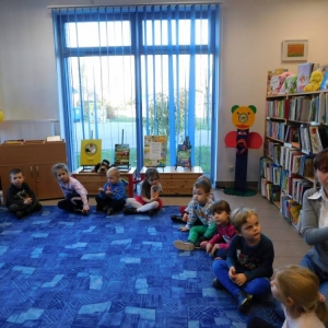 Dzieci siedzą na dywanie w kółeczku i słuchają pani bibliotekarki